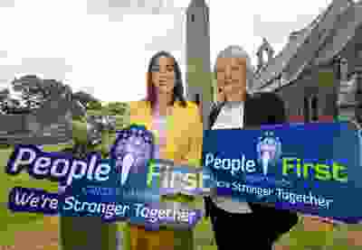 two women holding signs for People First Credit Union with the slogan "We're Stronger Together." They are standing outside in front of an old stone church and a round tower, suggesting a local or historical setting