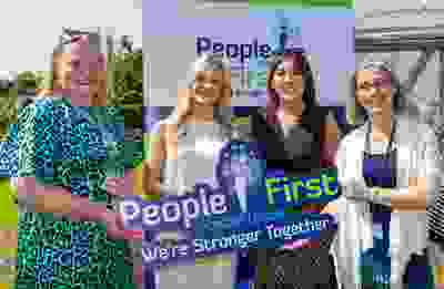 Four women smiling and holding a People First Credit Union sign that reads "We're Stronger Together" during a sunny event at Scoil Mhuire Abbeyleix. A People First Credit Union banner and a school polytunnel are visible in the background.